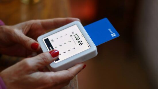 Close-up of a SumUp card reader in use, with a blue debit card inserted. The handheld device displays the transaction amount, showing how easy and efficient it is for small businesses to accept card payments. The user’s red nails contrast with the sleek white design of the card reader, capturing a modern, cashless payment moment.
